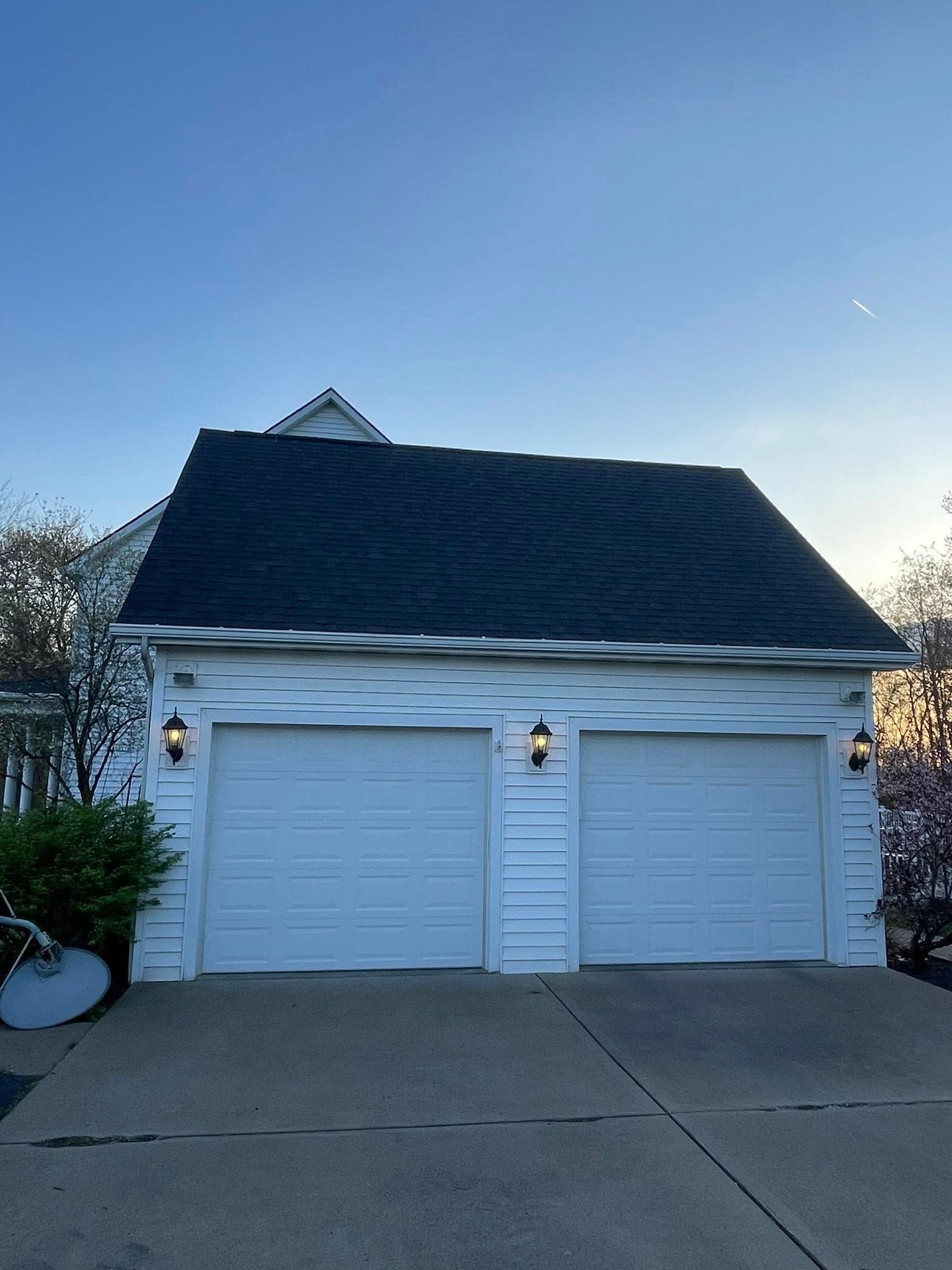 White two-car garage with a dark roof under a blue sky. Garage has two garage doors, lights, and a concrete driveway.