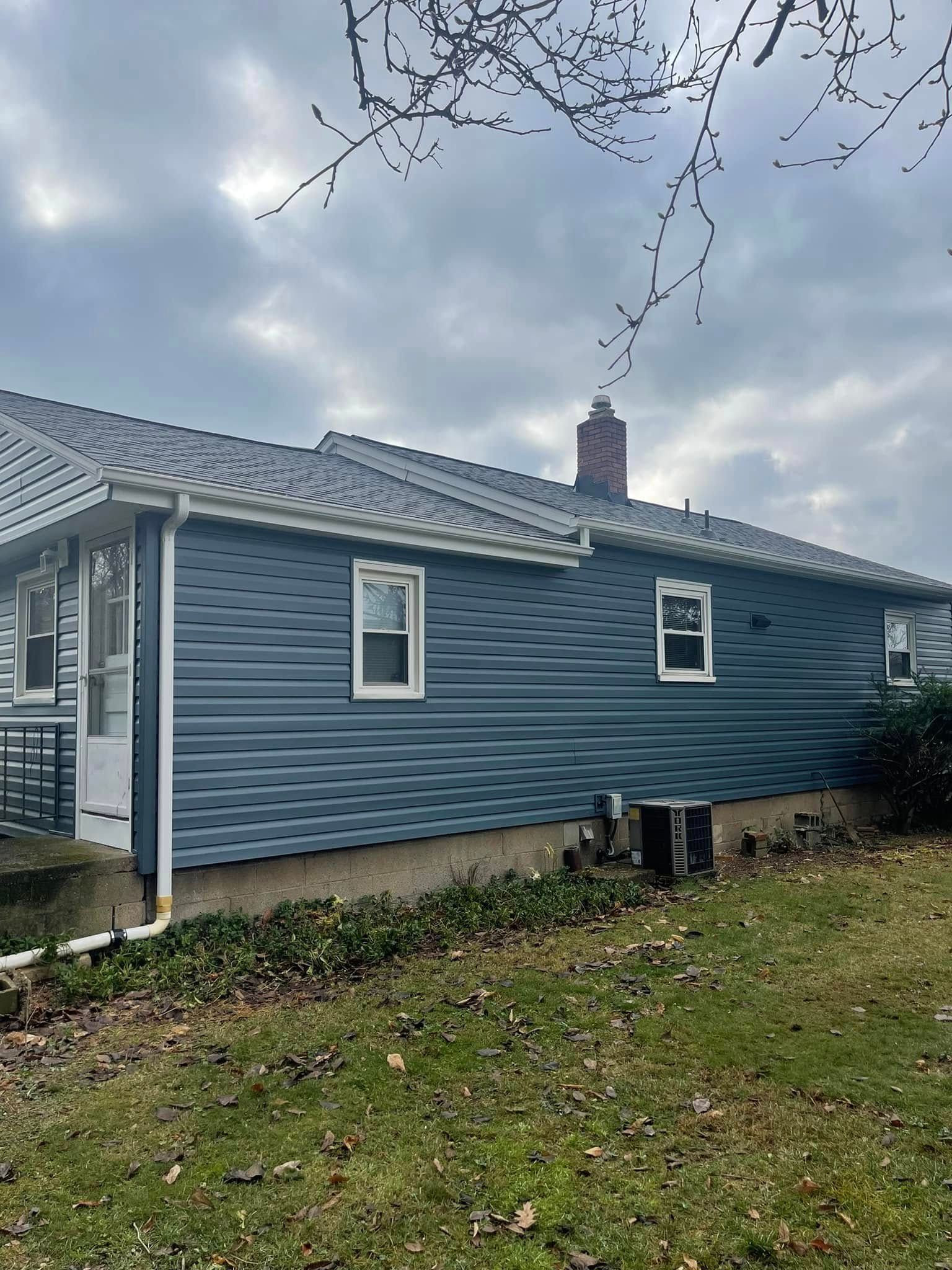 Blue-sided house with white-framed windows and a gray roof on a cloudy day. Brown grass and bare branches in the foreground.
