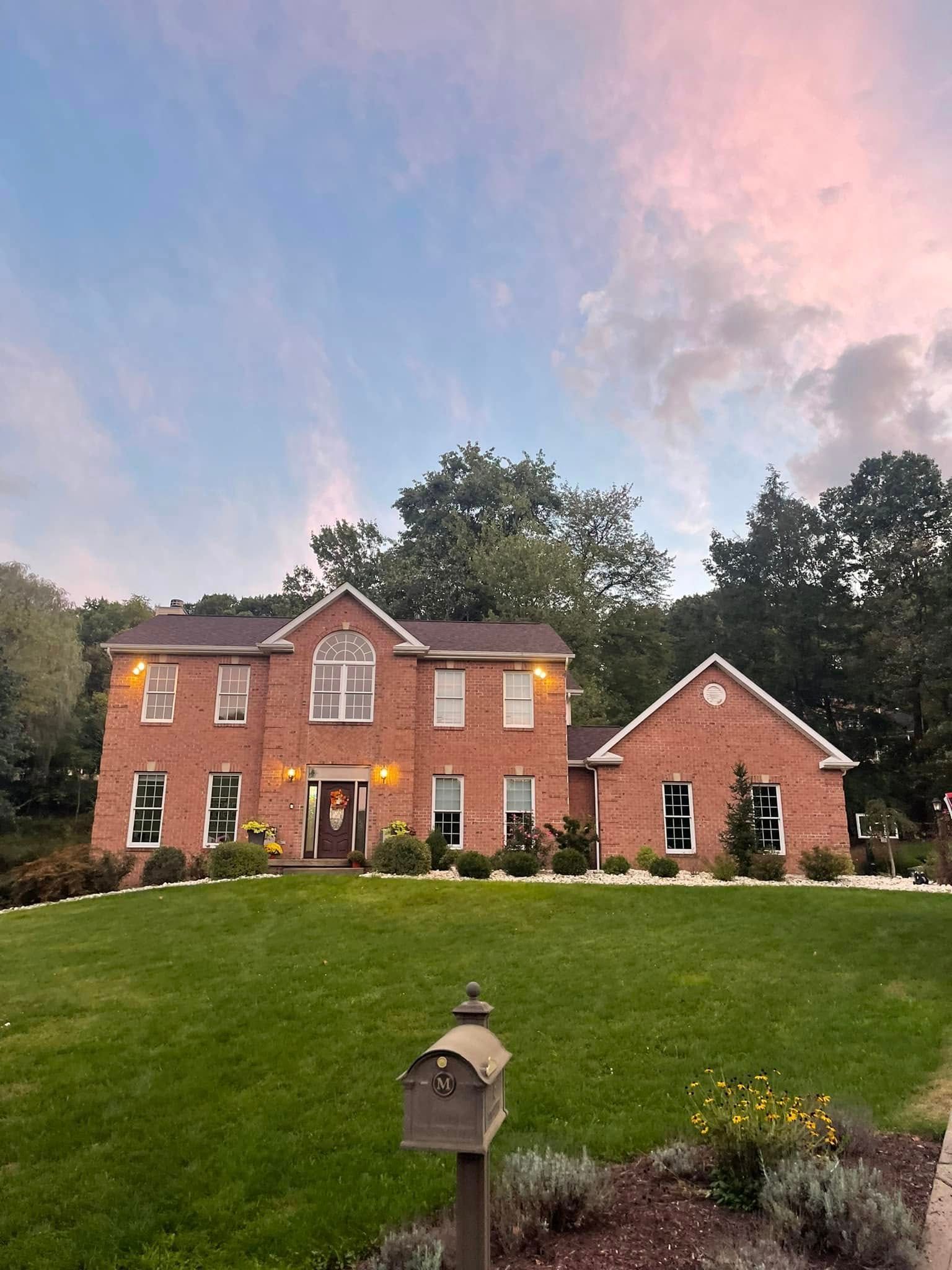 Two-story brick house with green lawn and dusk sky. A mailbox and flowers are in the foreground.