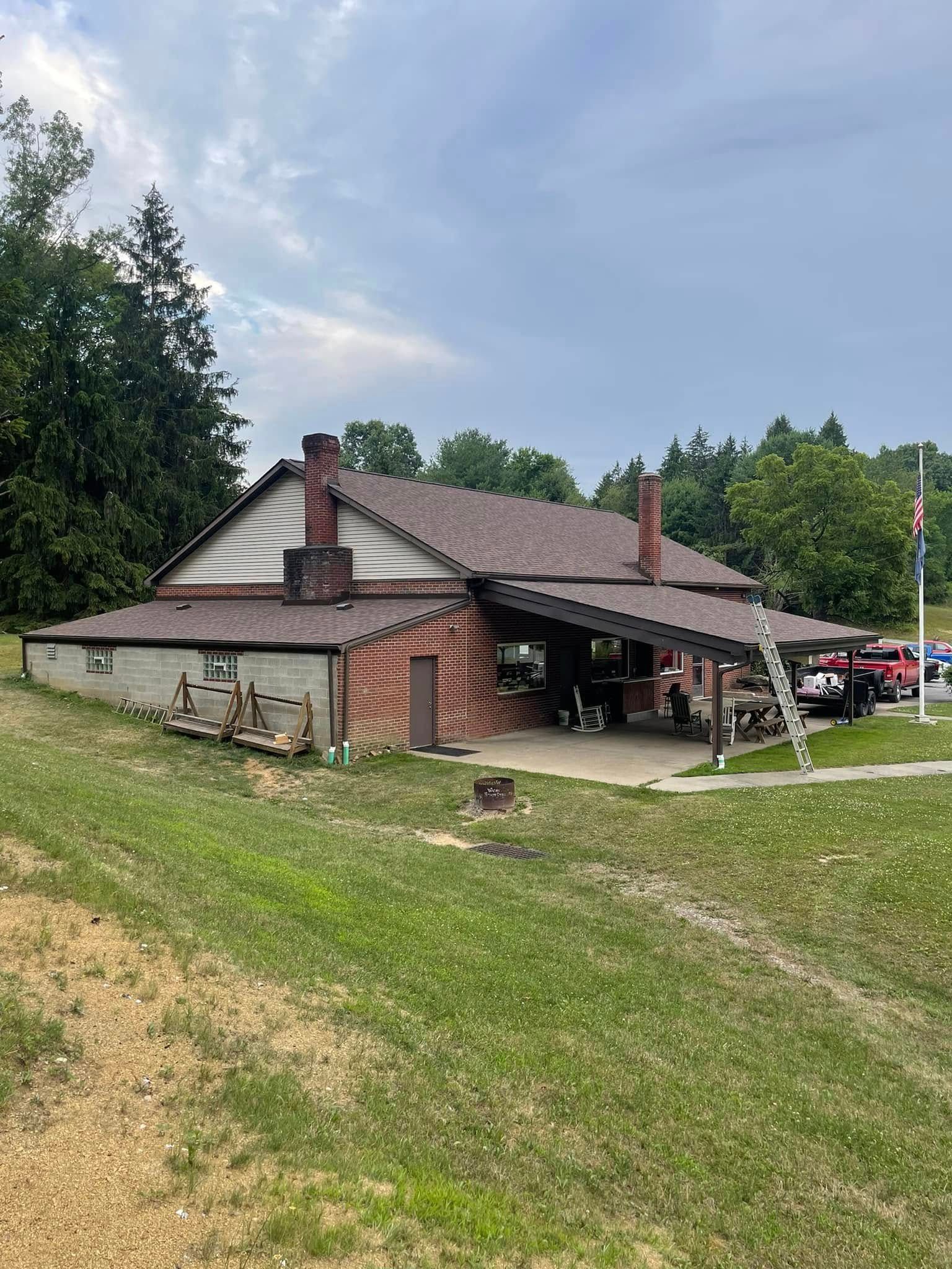 A brown brick and wood building with a covered patio, chimney, and new roof, set on a grassy hill under a cloudy sky.