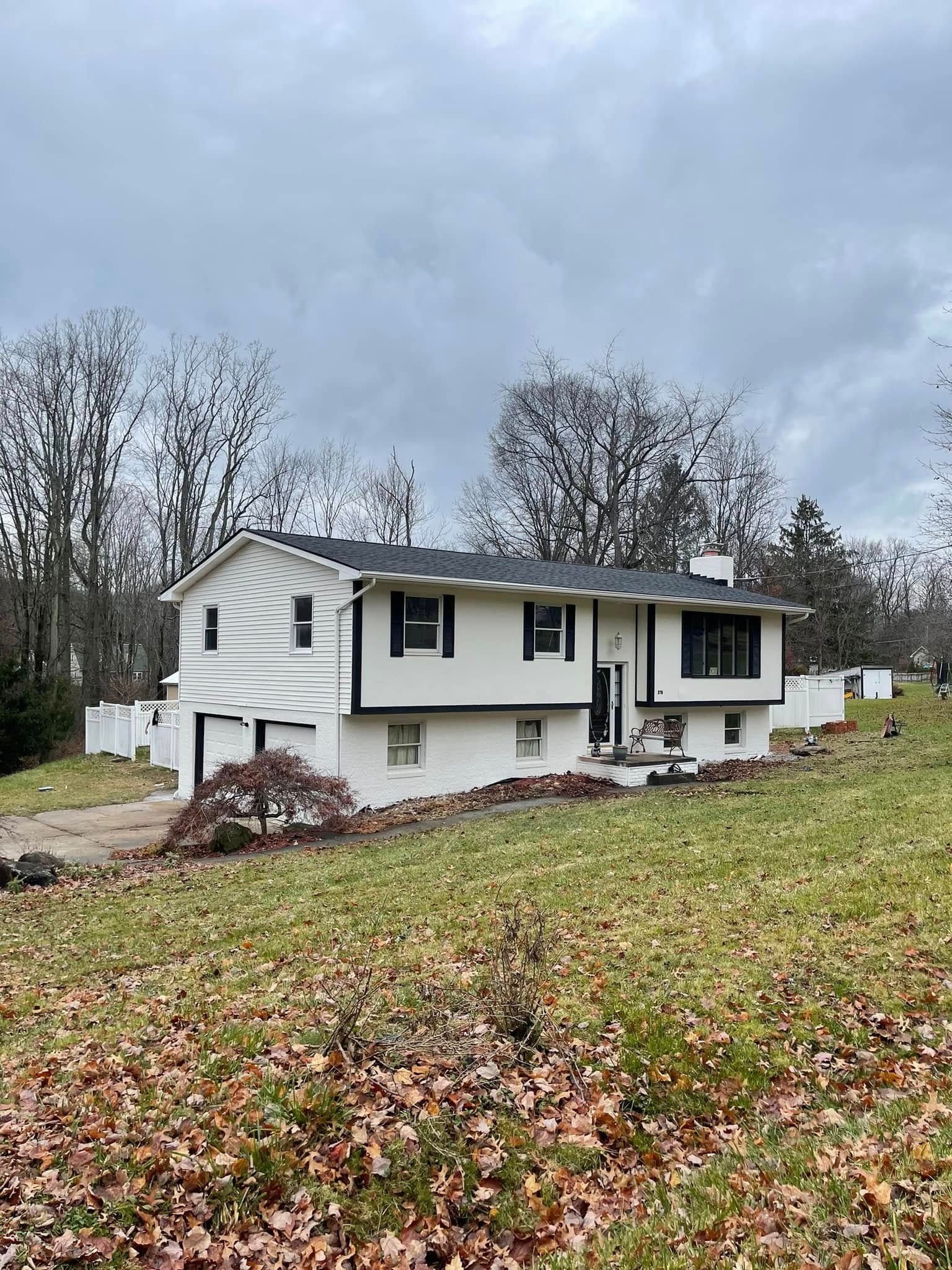 White two-story house on a hillside with a dark roof and trim. Overcast sky and bare trees in the background.