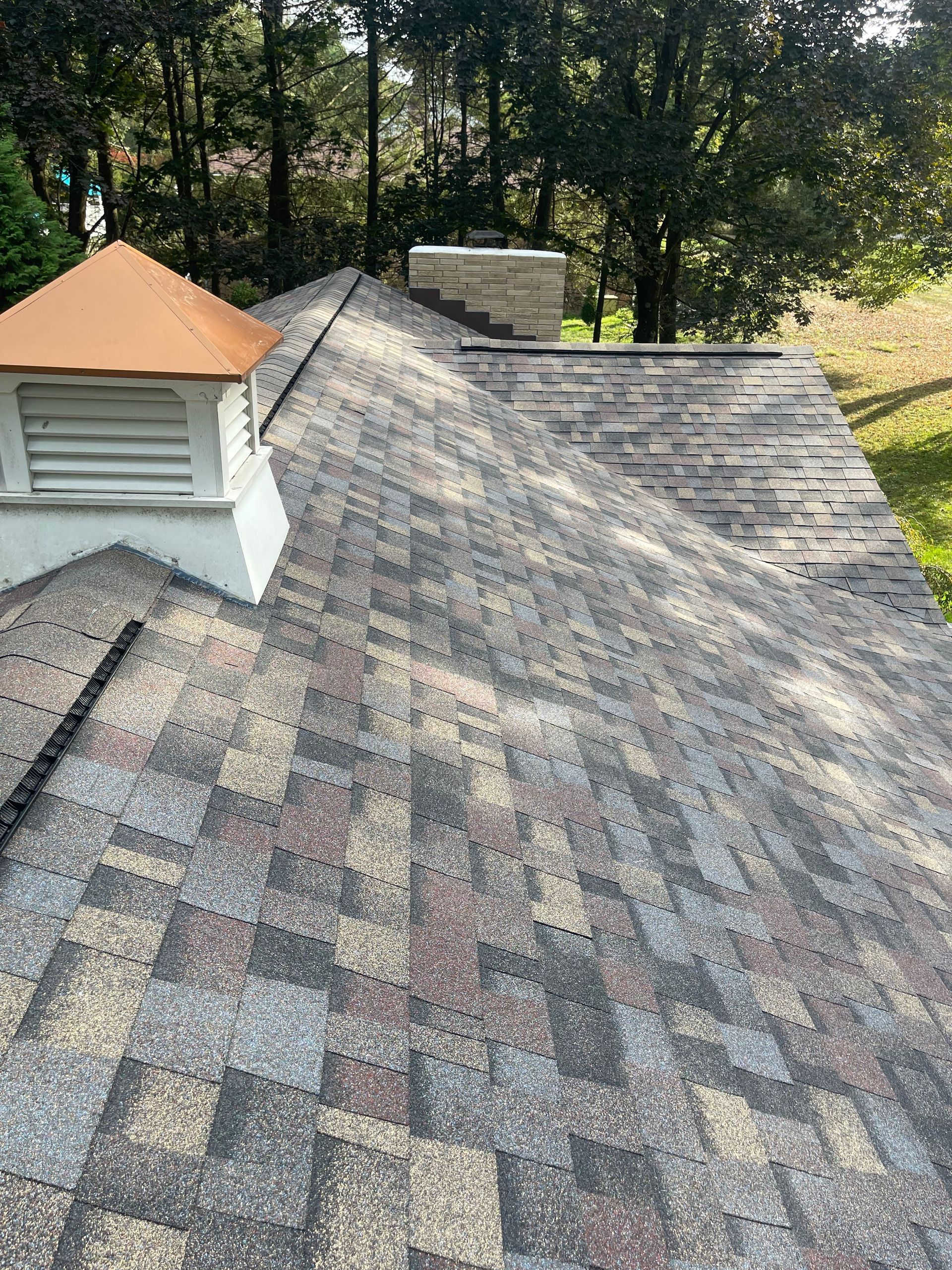 A roof with weathered shingles, a white cupola, and a brick chimney against a green trees.