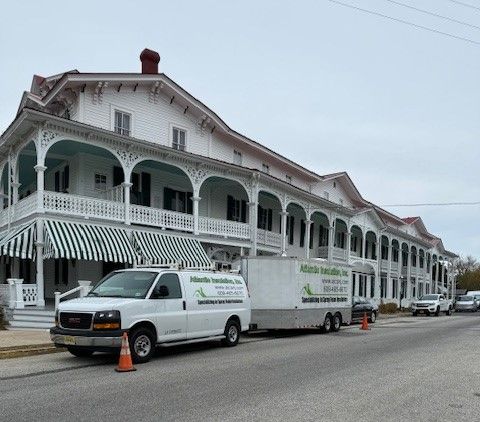 White building with porch, vehicles parked in front, including a service van and trailer.