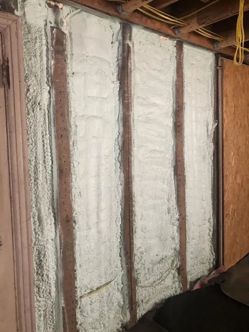 White spray foam insulation fills wall studs, brown wood visible. Wiring runs above.