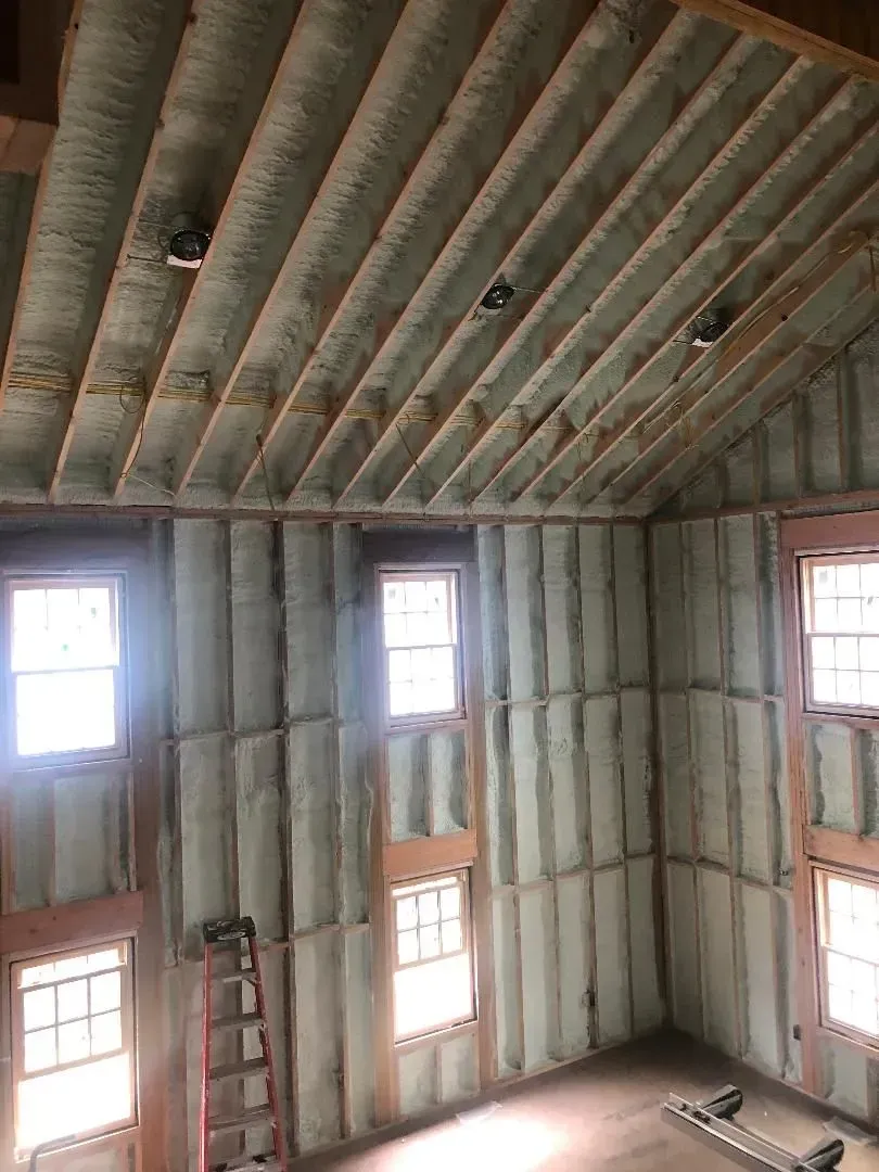 Interior view of a building under construction, showing spray foam insulation on walls and ceiling, with windows.