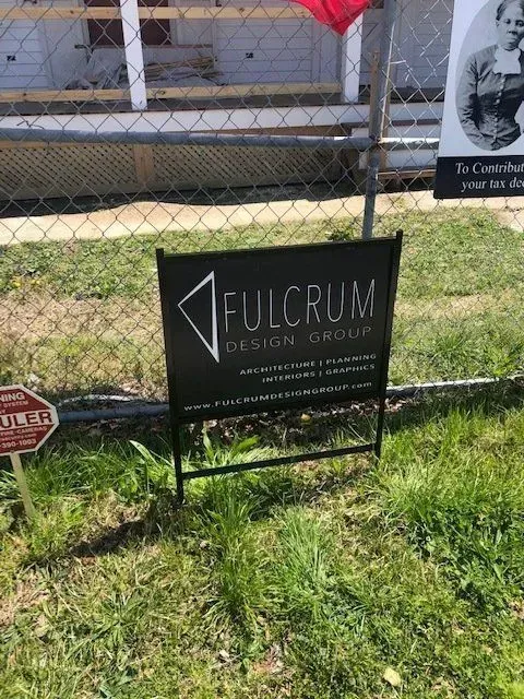 Sign for Fulcrum Design Group on grass, with fence and building in background.