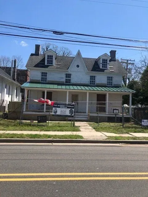 Two-story white house with green roof, porch, and dormers, behind a sidewalk and road under a blue sky.