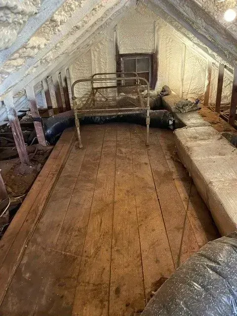 Attic with wooden floorboards, a metal bed frame, small window, and insulation.