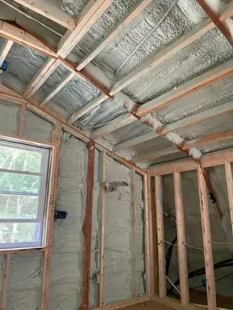 Interior view of a room with exposed wooden framing and spray foam insulation on walls and ceiling. A window is visible.