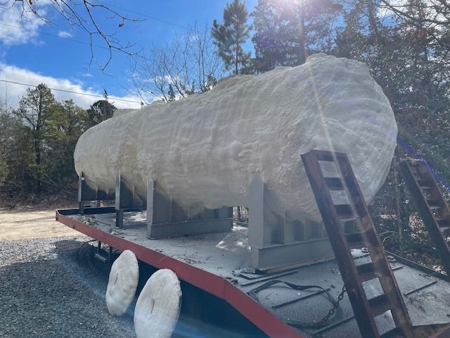 Large, foam-covered tank on a flatbed trailer, outdoors. Blue sky, trees in the background.