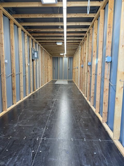 Narrow unfinished hallway with exposed wooden studs, black floor, and ceiling lights.