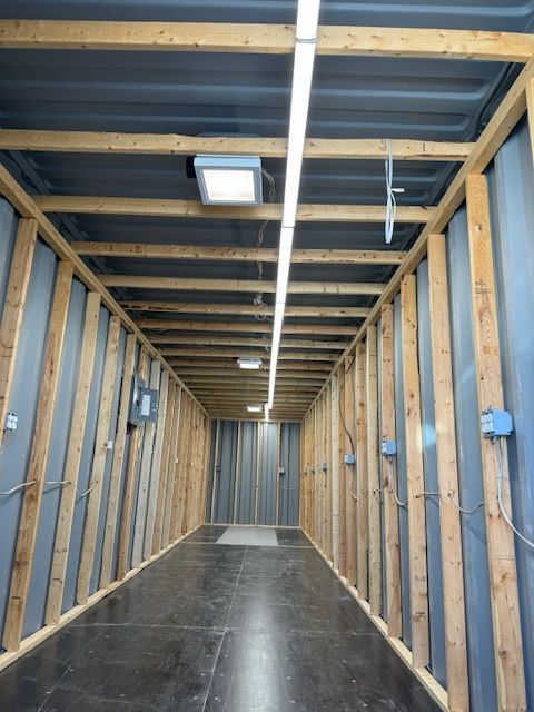 Empty unfinished hallway with exposed wooden framing and a concrete floor, lit by a ceiling strip light.
