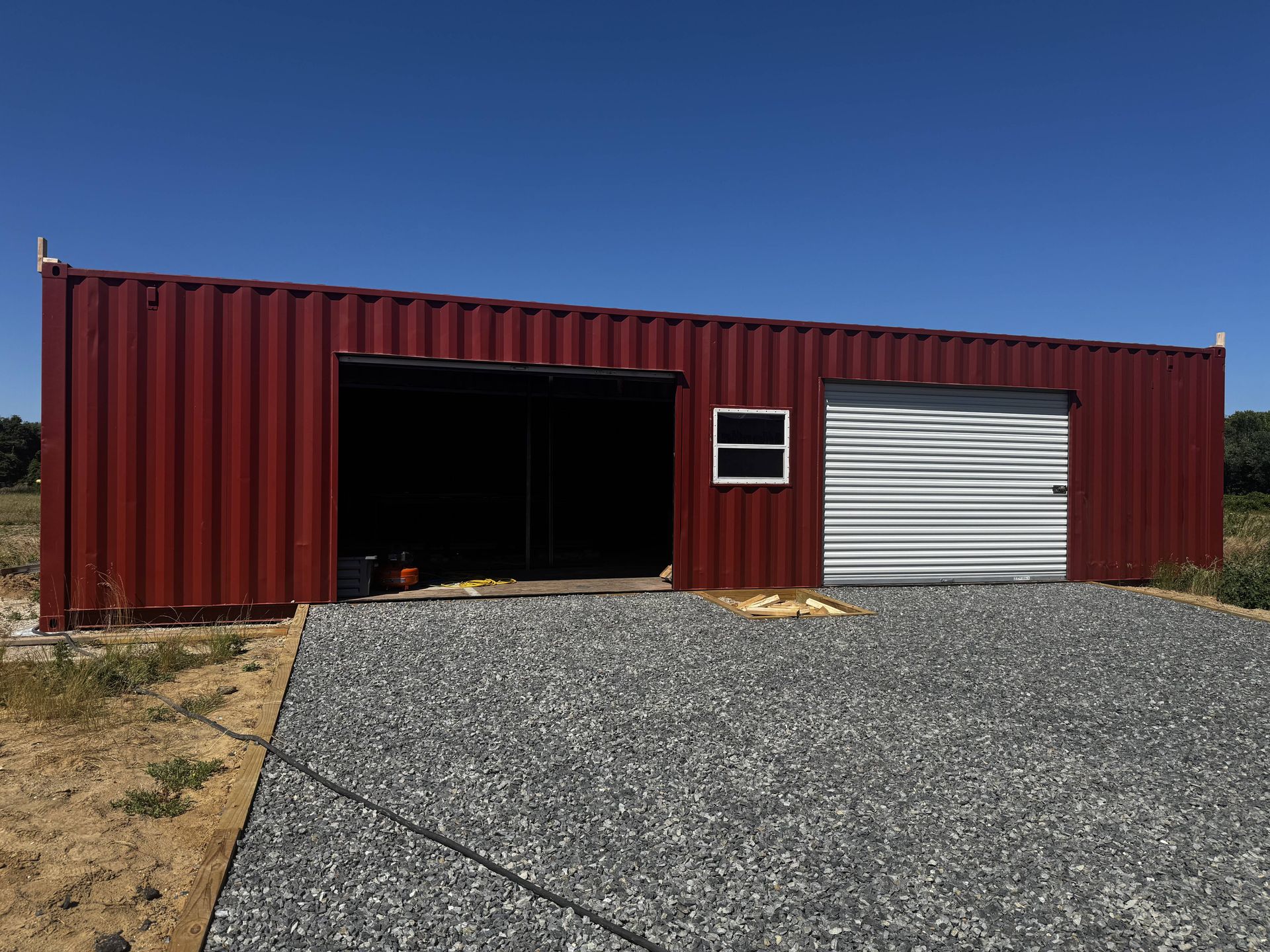 Red metal storage building with a large open bay and a white roll-up door on gravel under a blue sky