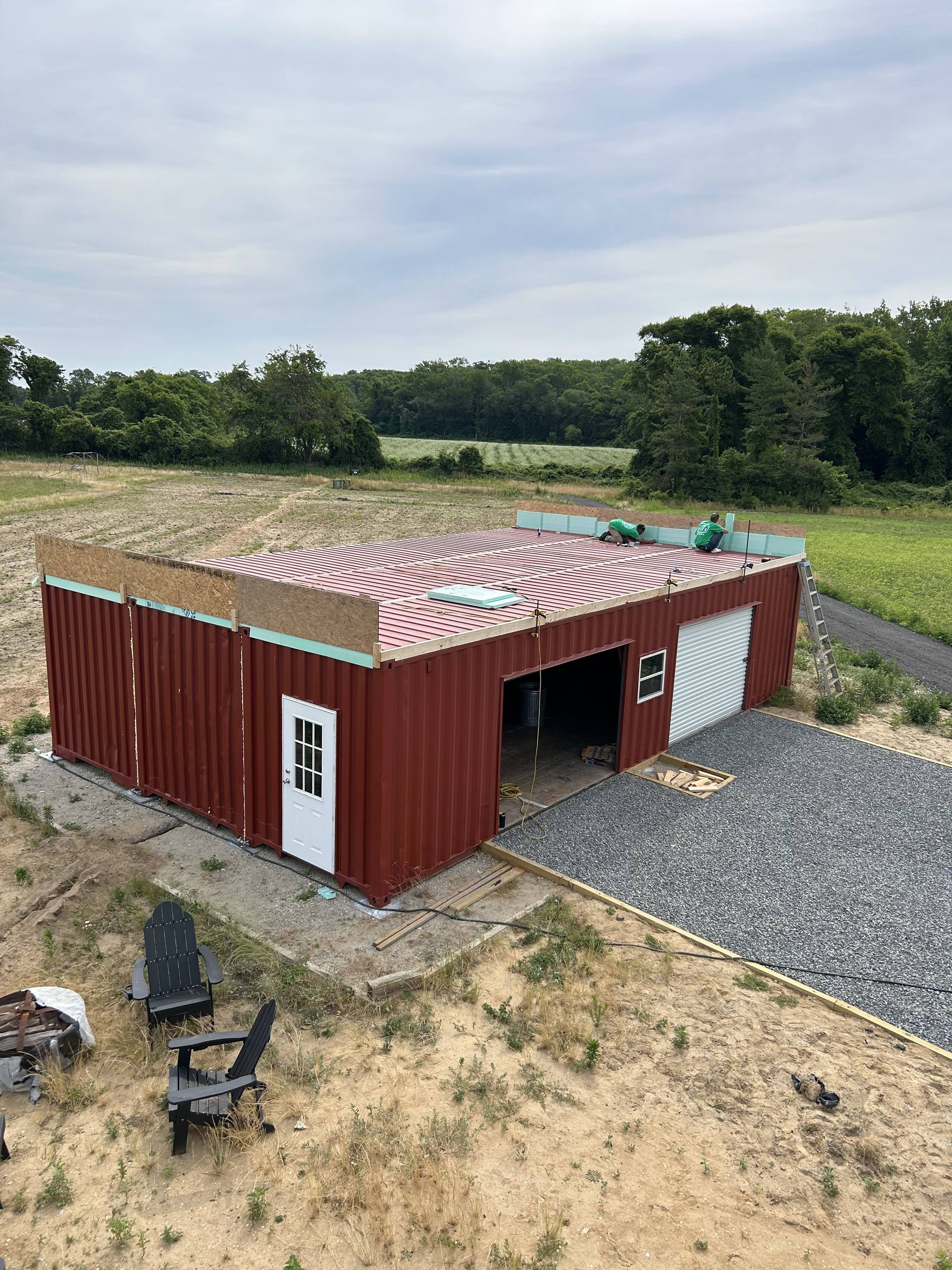 Red shipping container shed with patterned roof on gravel in a rural field, with lawn chairs nearby.
