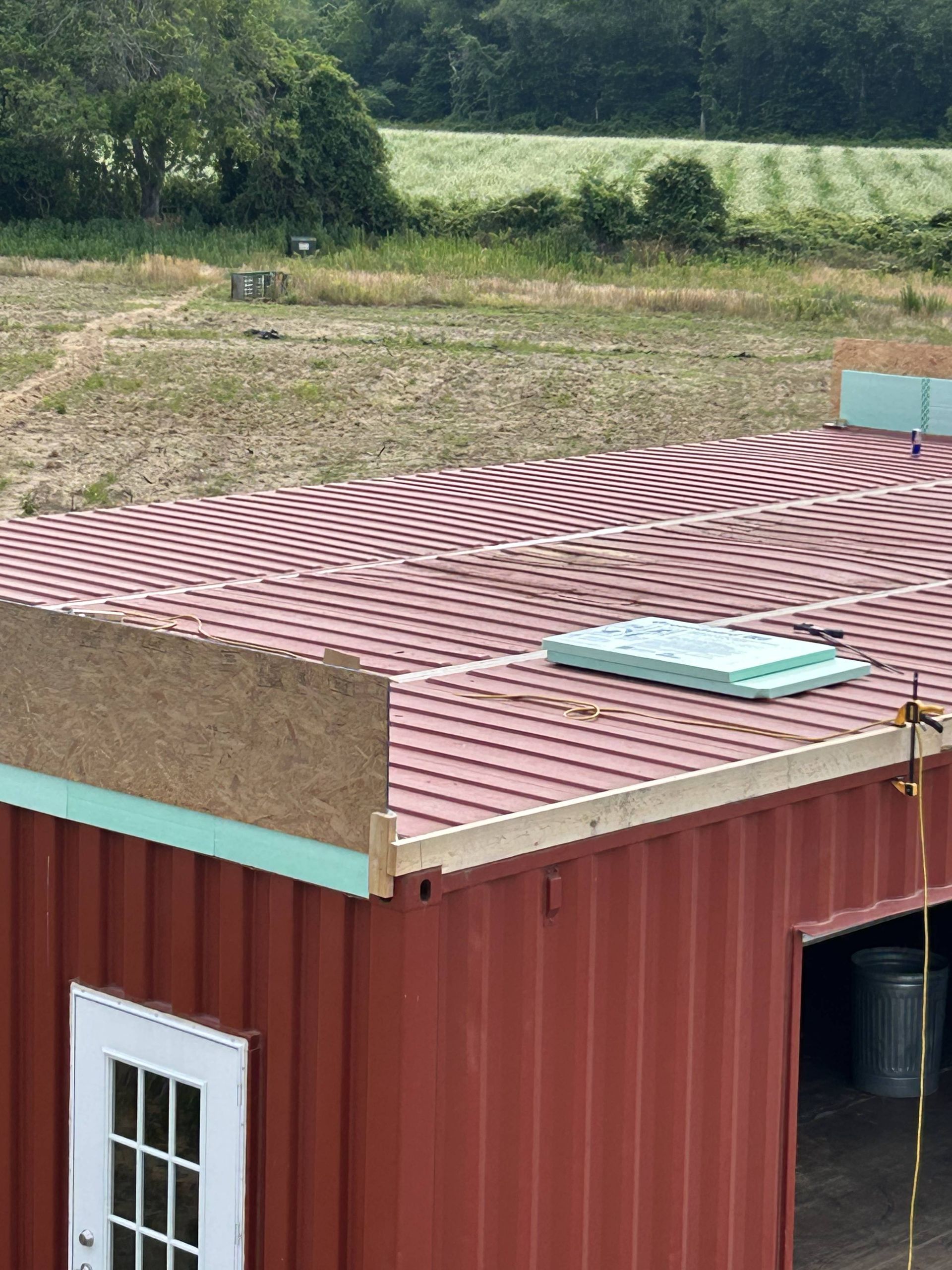 Red rooftop of a building with a skylight, set beside grassy fields and trees.