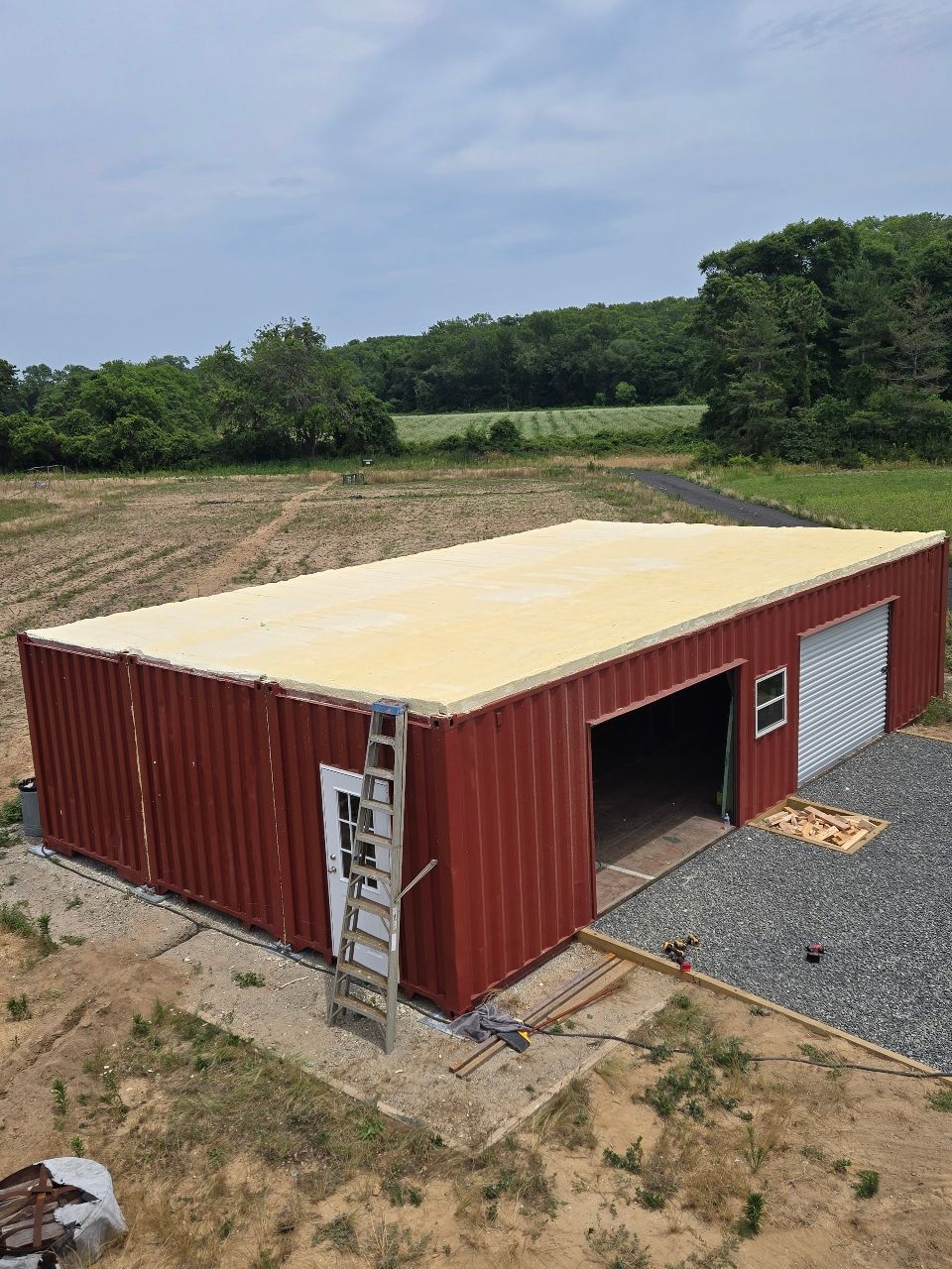 Red shipping-container workshop with open bay door, ladder, and gravel pad in a rural field.
