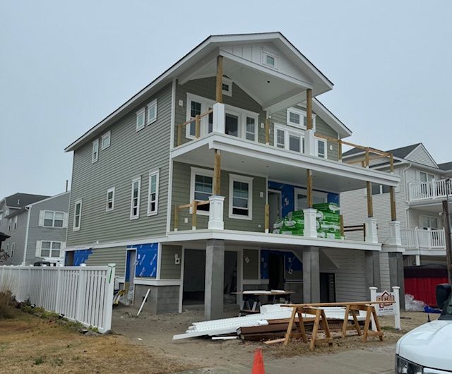 Two-story house under construction with a porch and gray siding. Lumber and building materials are present.