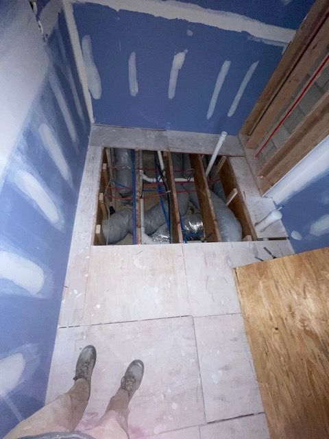 Bathroom under construction, with exposed plumbing and plywood flooring. Drywall on walls and ceiling; legs visible.