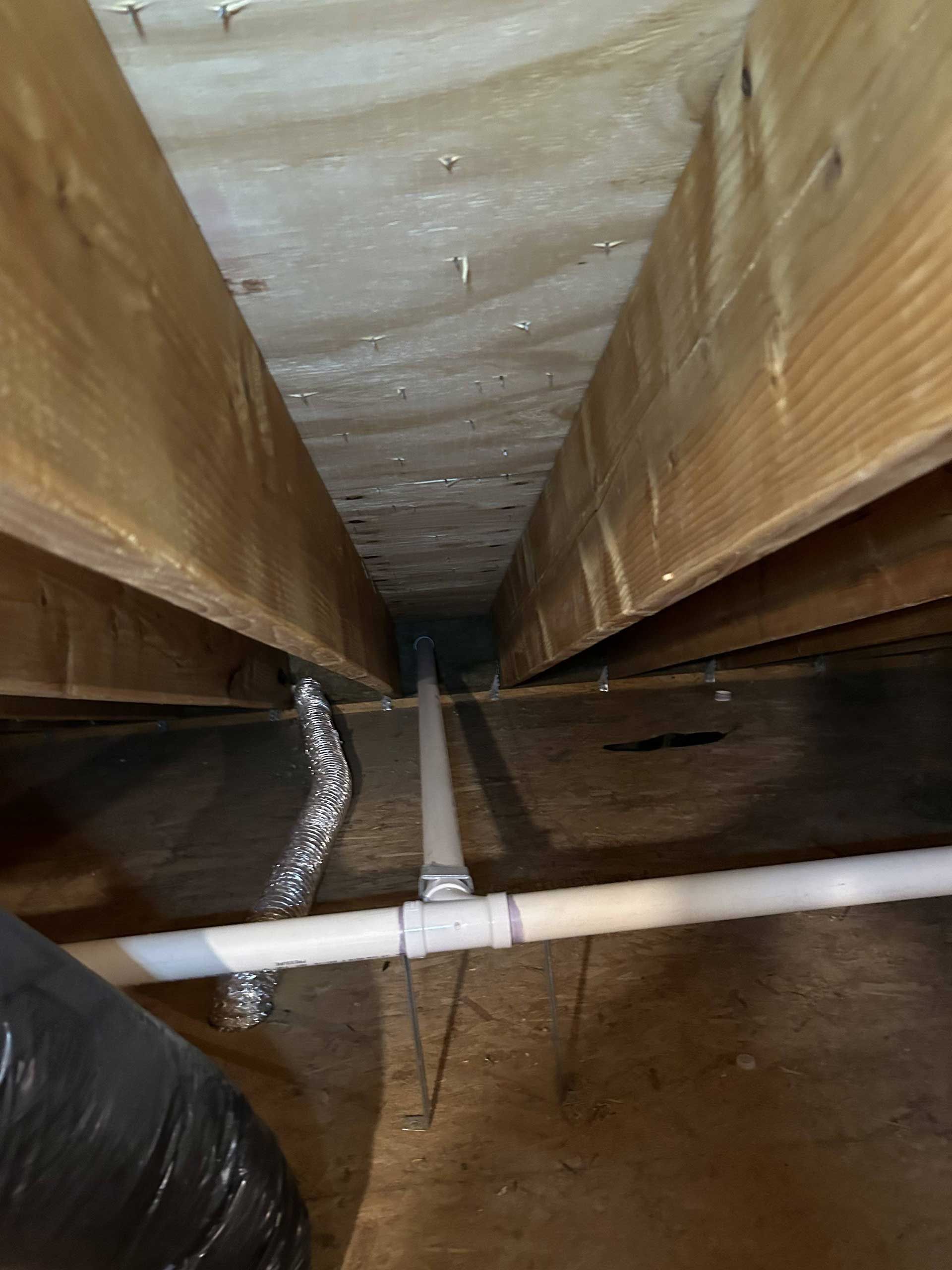 A view between wooden floor joists showing white PVC pipes, a silver flexible duct, and dark floor insulation.