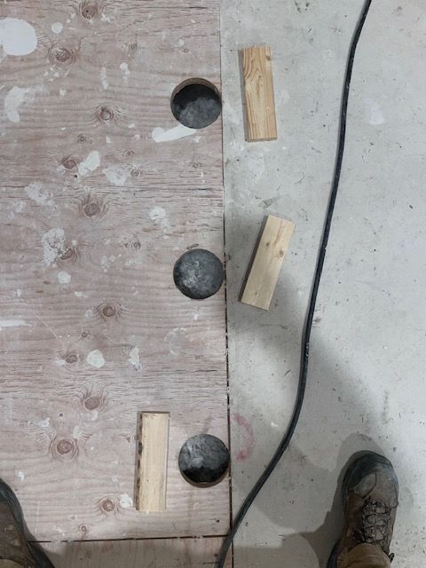 Wooden floorboard with three circular holes and wooden blocks next to a concrete floor. A black cable and work boots are visible.