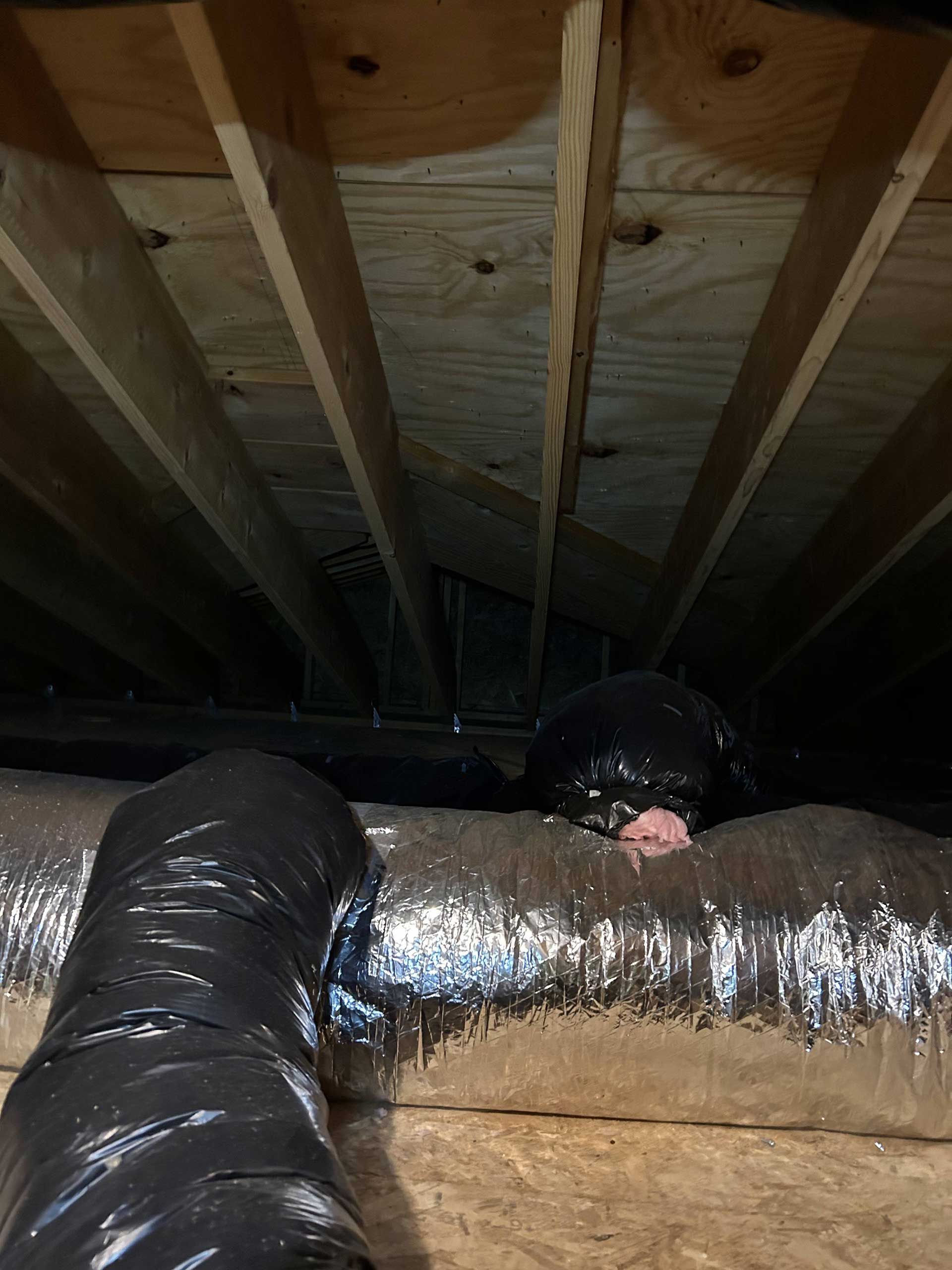 Attic view showing flexible ductwork connected to a rigid vent, with plywood floor joists above.