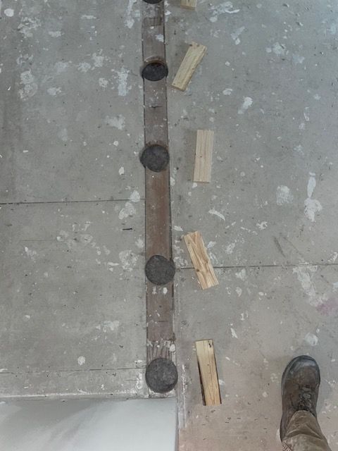 Strip of metal with round, dark plugs, positioned on concrete floor with wood shims scattered around it, foot at bottom.