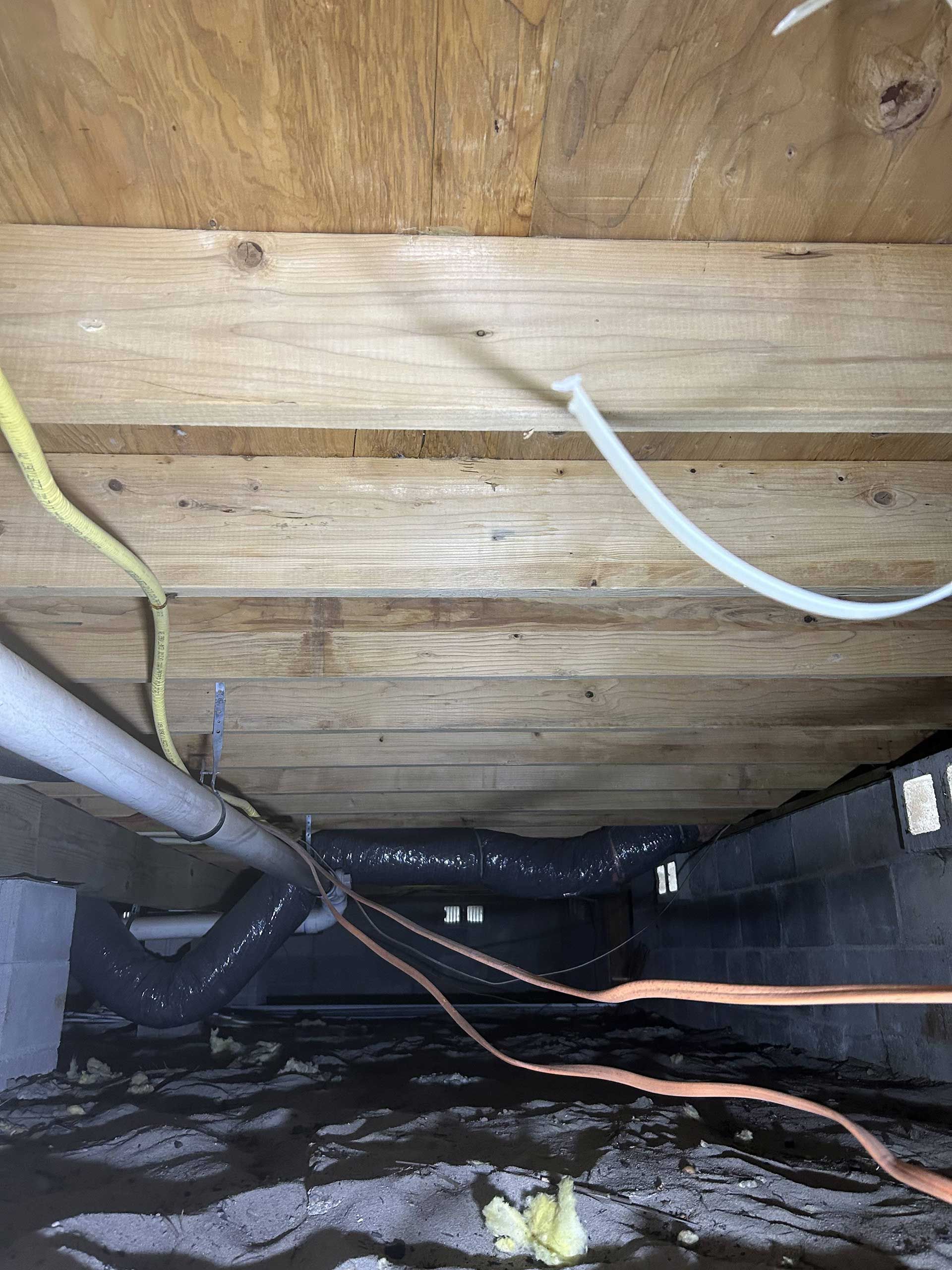 View under a wooden floor showing floor joists, electrical wiring, ductwork, and dark, damp soil in a crawl space.