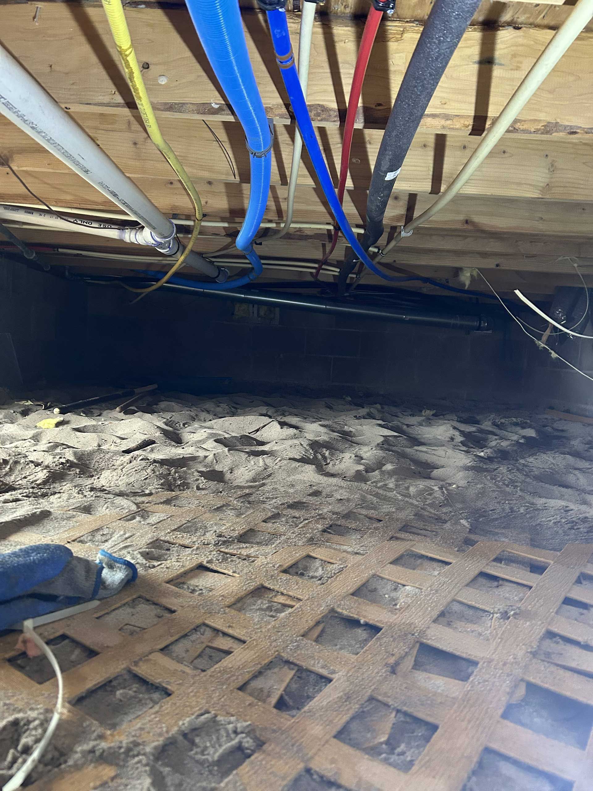 A crawl space with exposed wooden joists, multicolored PEX plumbing pipes, and a dirt floor covered in a grid mat.