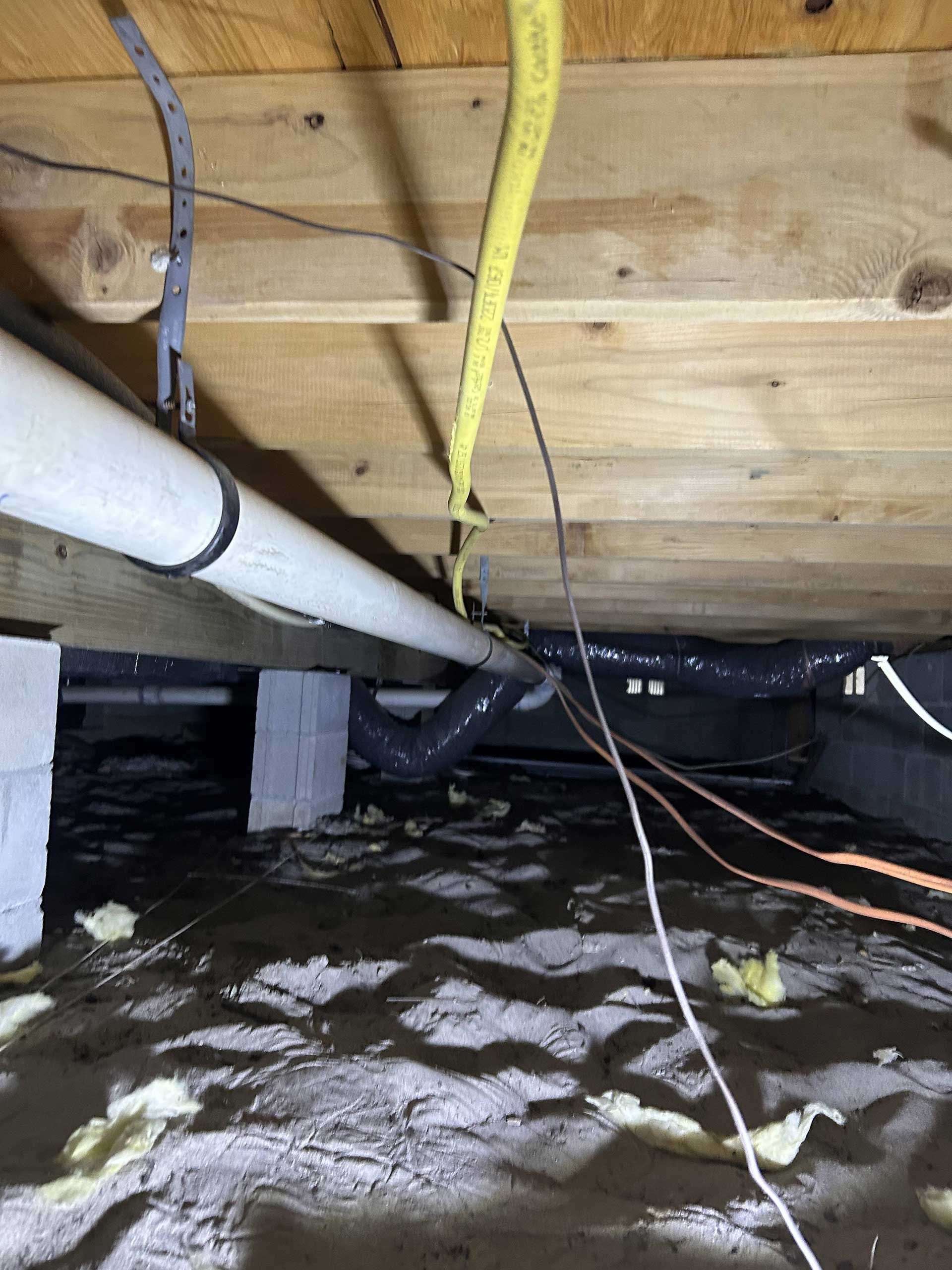 A crawl space with wooden floor joists, a white plumbing pipe, a yellow gas line, and exposed insulation on the ground.