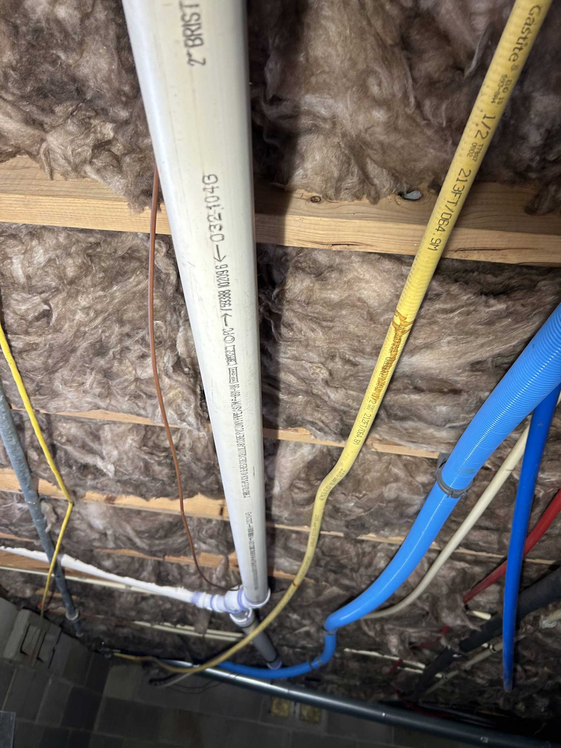 Plumbing pipes and electrical wiring exposed against fiberglass insulation in an unfinished ceiling space.