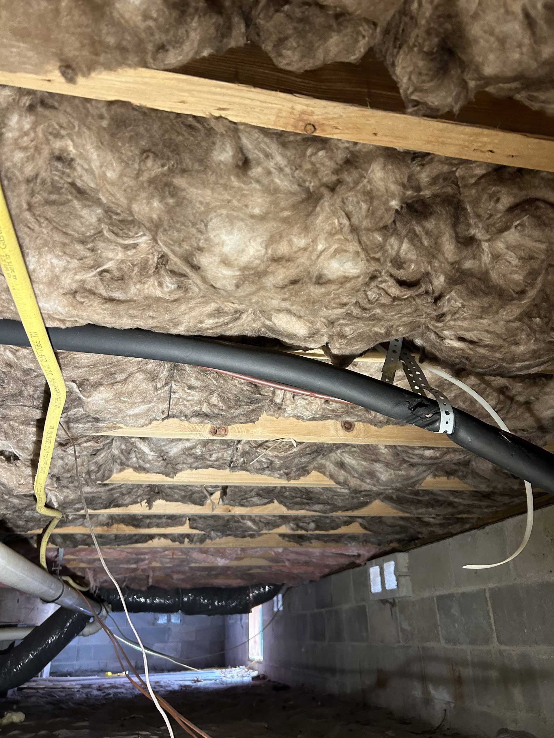 A view of fiberglass insulation installed between wooden floor joists in a crawlspace with various utility pipes and wires.