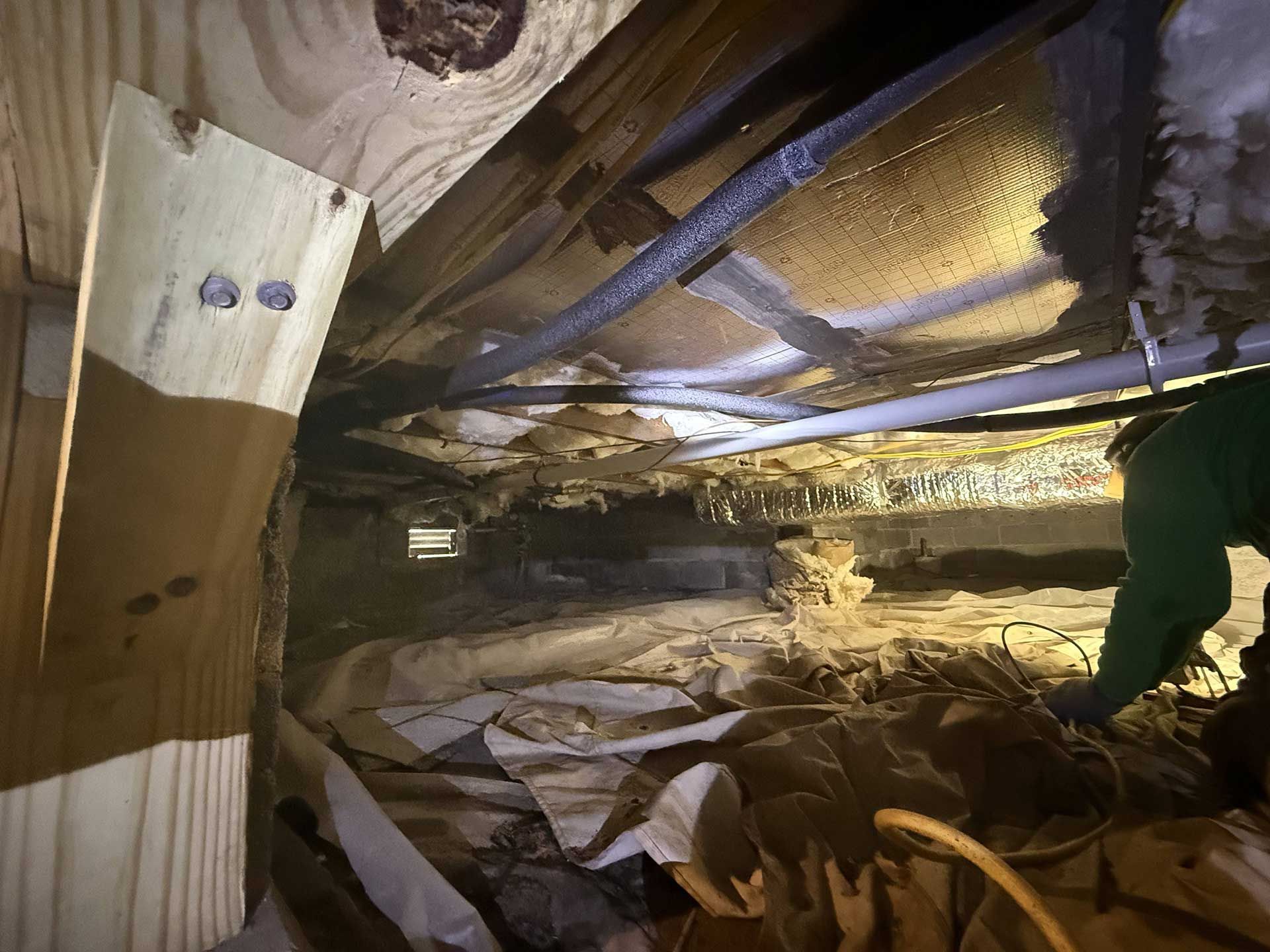 A crawl space showing fiberglass insulation installed between wooden floor joists, with pipes and wiring visible below.