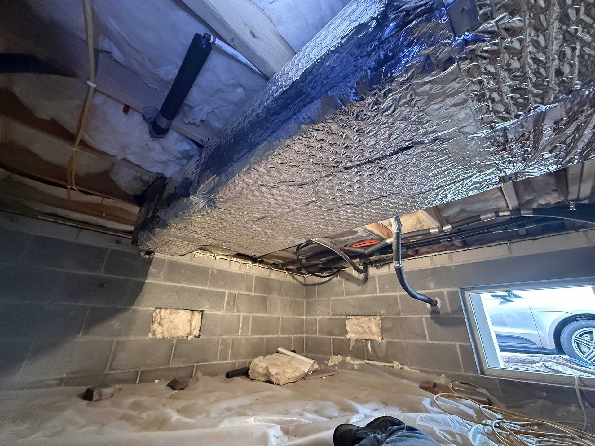A low-angle view of a crawl space with insulated ducts, exposed wooden floor joists, concrete blocks, and a foundation vent.
