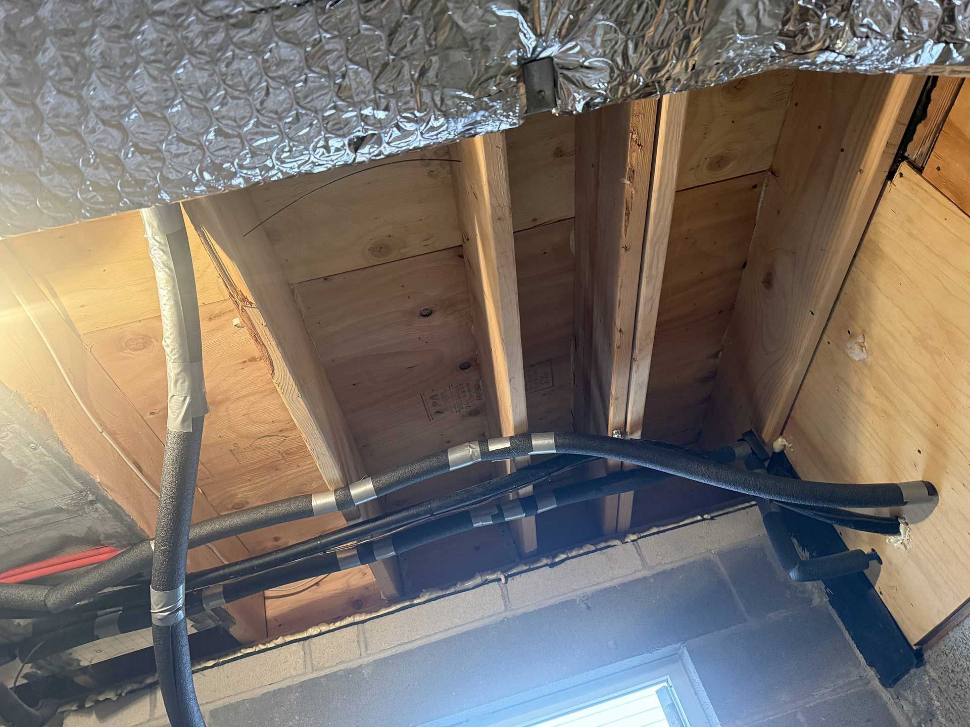 Exposed wooden floor joists and subflooring in a basement, with dark flexible hoses running across the ceiling area.