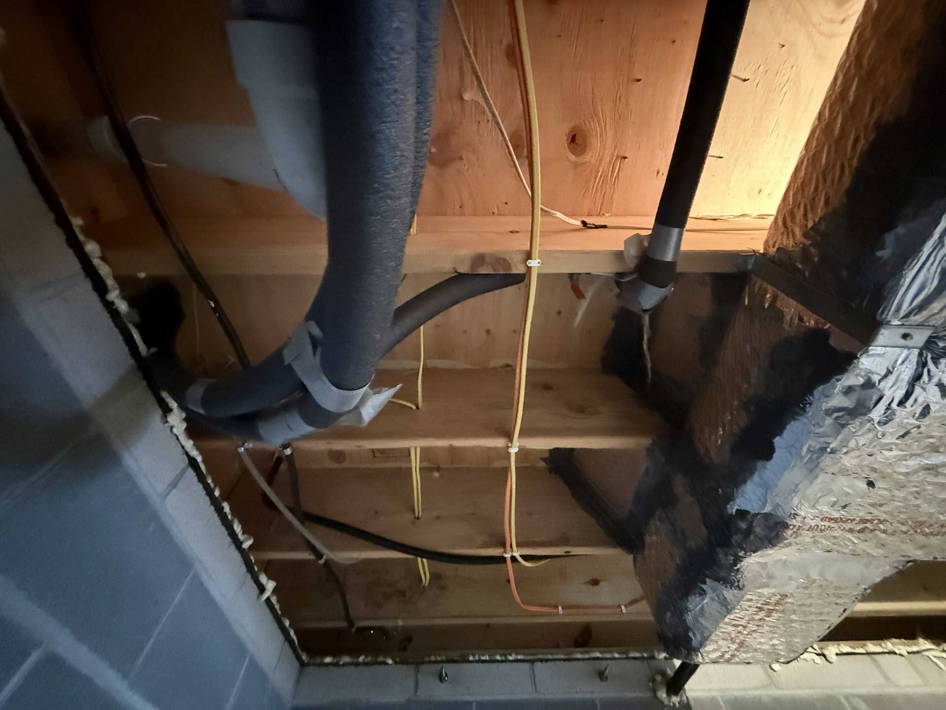Exposed wooden ceiling joists with insulated pipes and electrical wires running through the open floor framing.