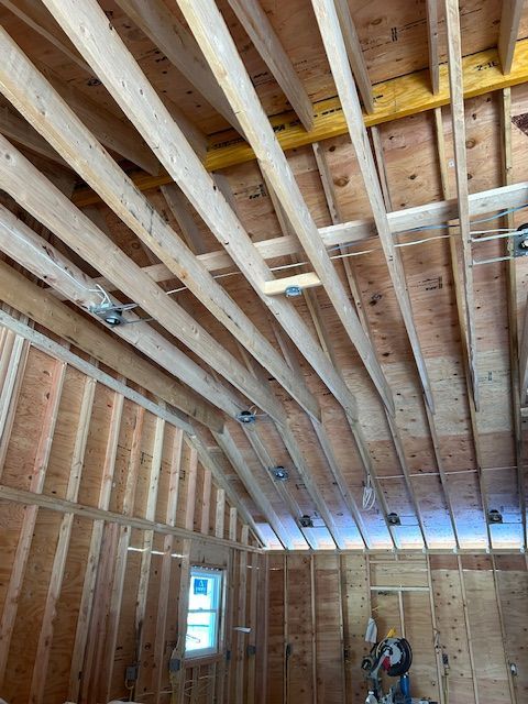 Interior view of a building under construction. Wooden rafters and studs are visible, with electrical boxes in the ceiling.