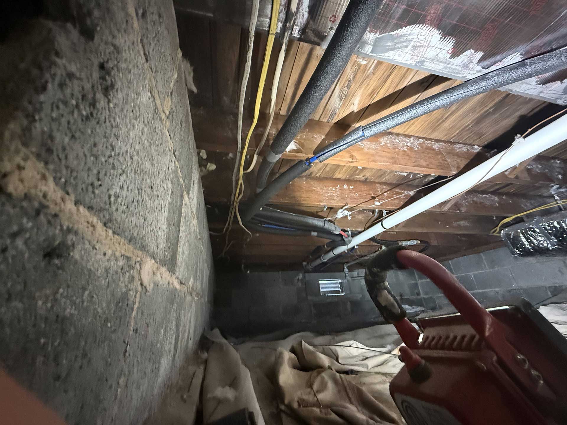 Crawl space view showing wooden joists, electrical wiring, pipes, and a block foundation wall, with a red tool in the corner.