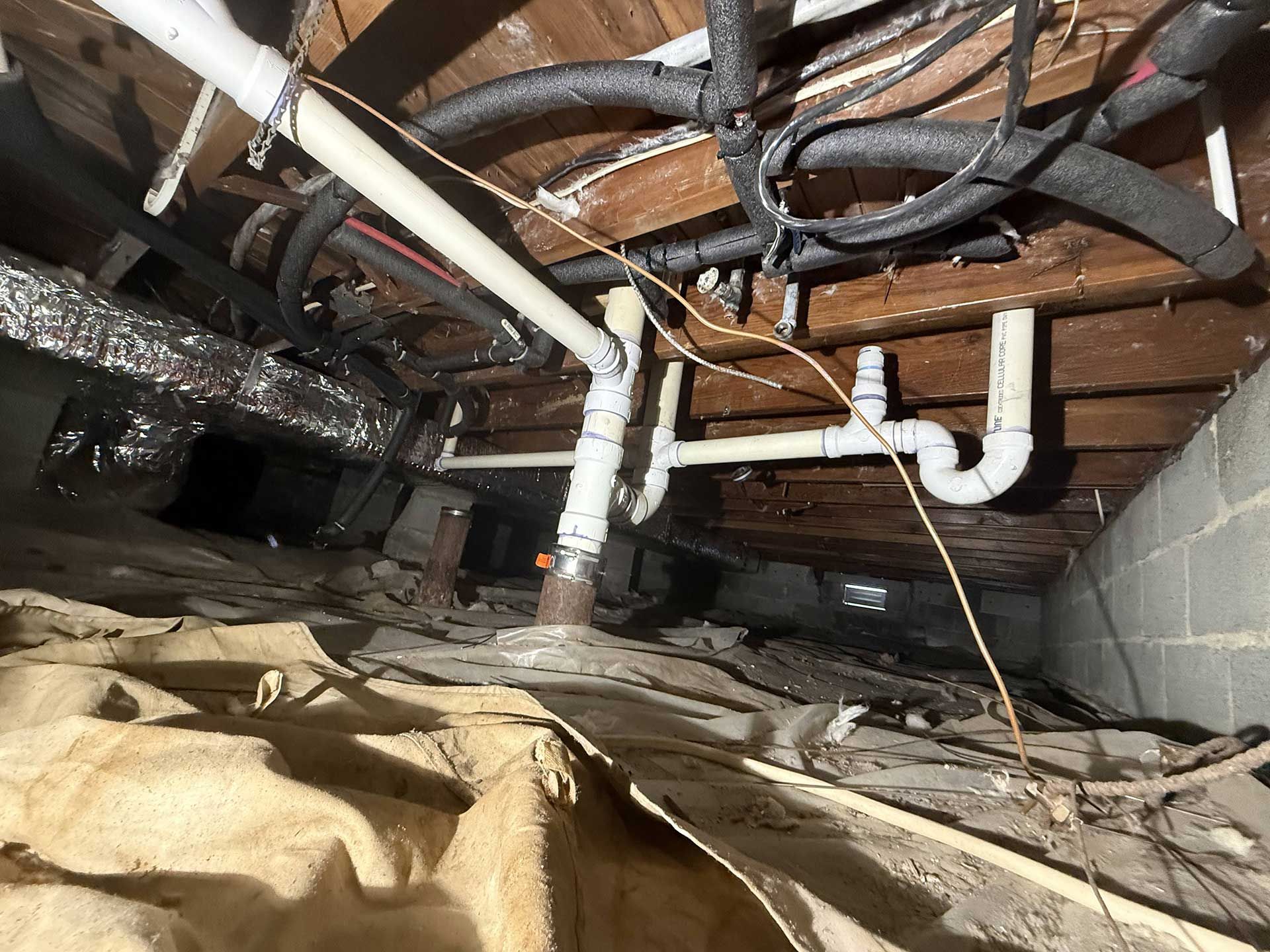 A crawl space containing white PVC plumbing pipes, black insulated wires, and a dirt floor with a plastic vapor barrier.