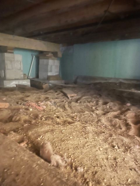 Crawl space with dirt floor, concrete supports, wooden beams above, and a light blue wall in the background.