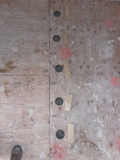 Plywood floor with several dark holes and wood shims. Red markings are present.