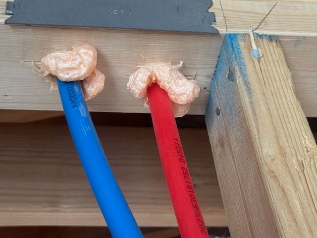 Blue and red PEX pipes pass through wood framing, sealed with pink foam.