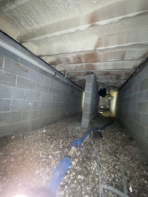 Low crawlspace with cinder block walls, insulation overhead, gravel floor, and blue tubing running through the center