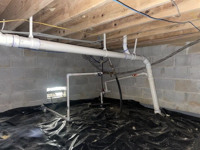 Crawl space with exposed pipes, cinder block walls, wooden joists, and black vapor barrier on the floor