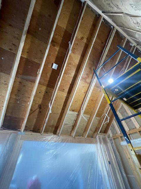 Interior under construction with exposed wooden rafters, plastic sheeting, and a ladder under bright work lights