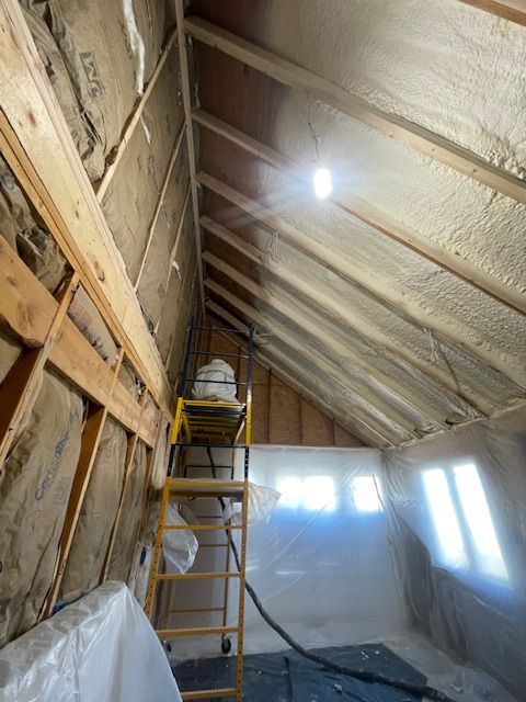 Insulated attic under renovation with ladder, exposed beams, and light through plastic-covered windows