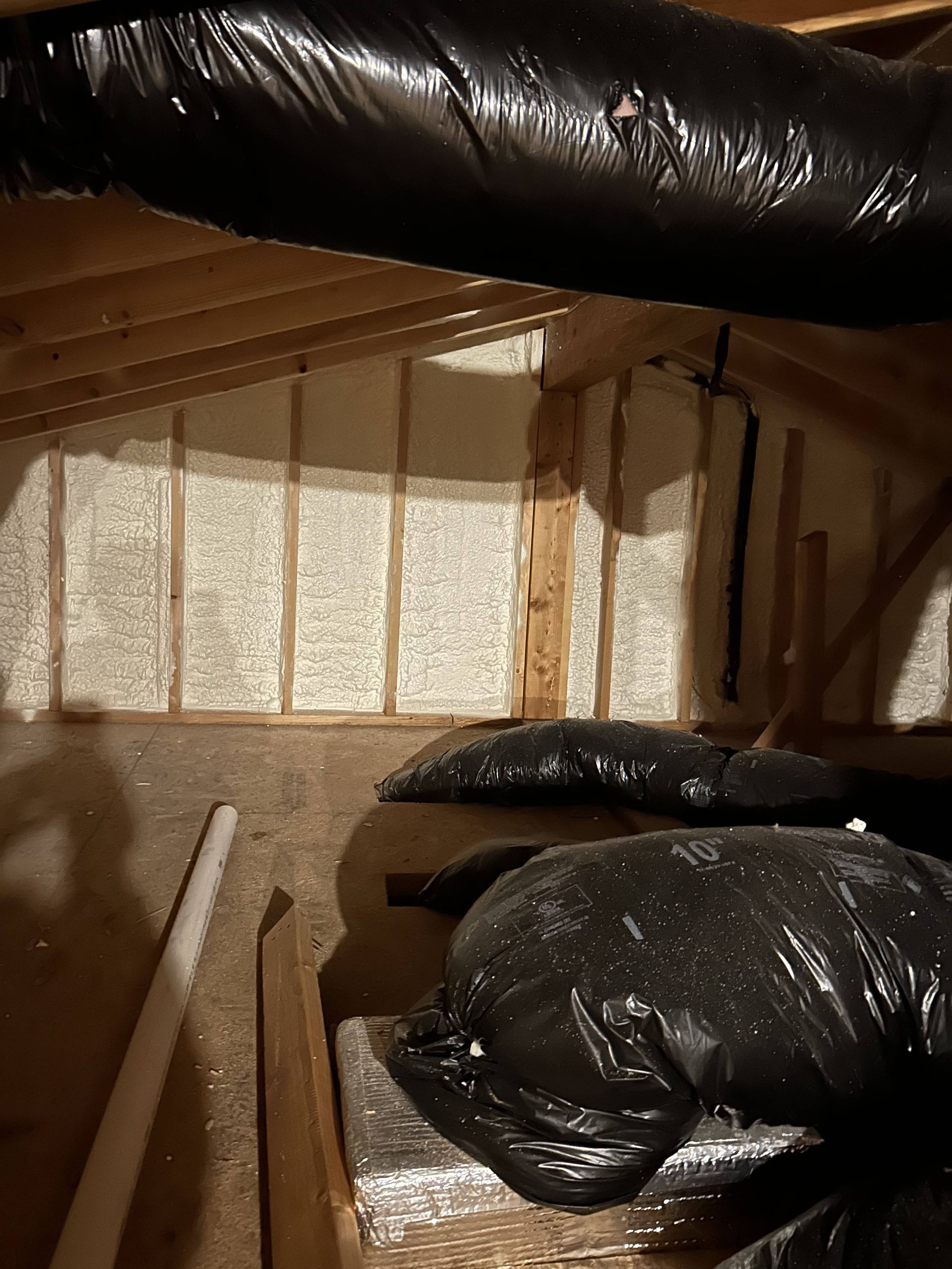 Attic crawlspace with black insulated ducting, exposed beams, and a white brick wall behind plastic sheeting