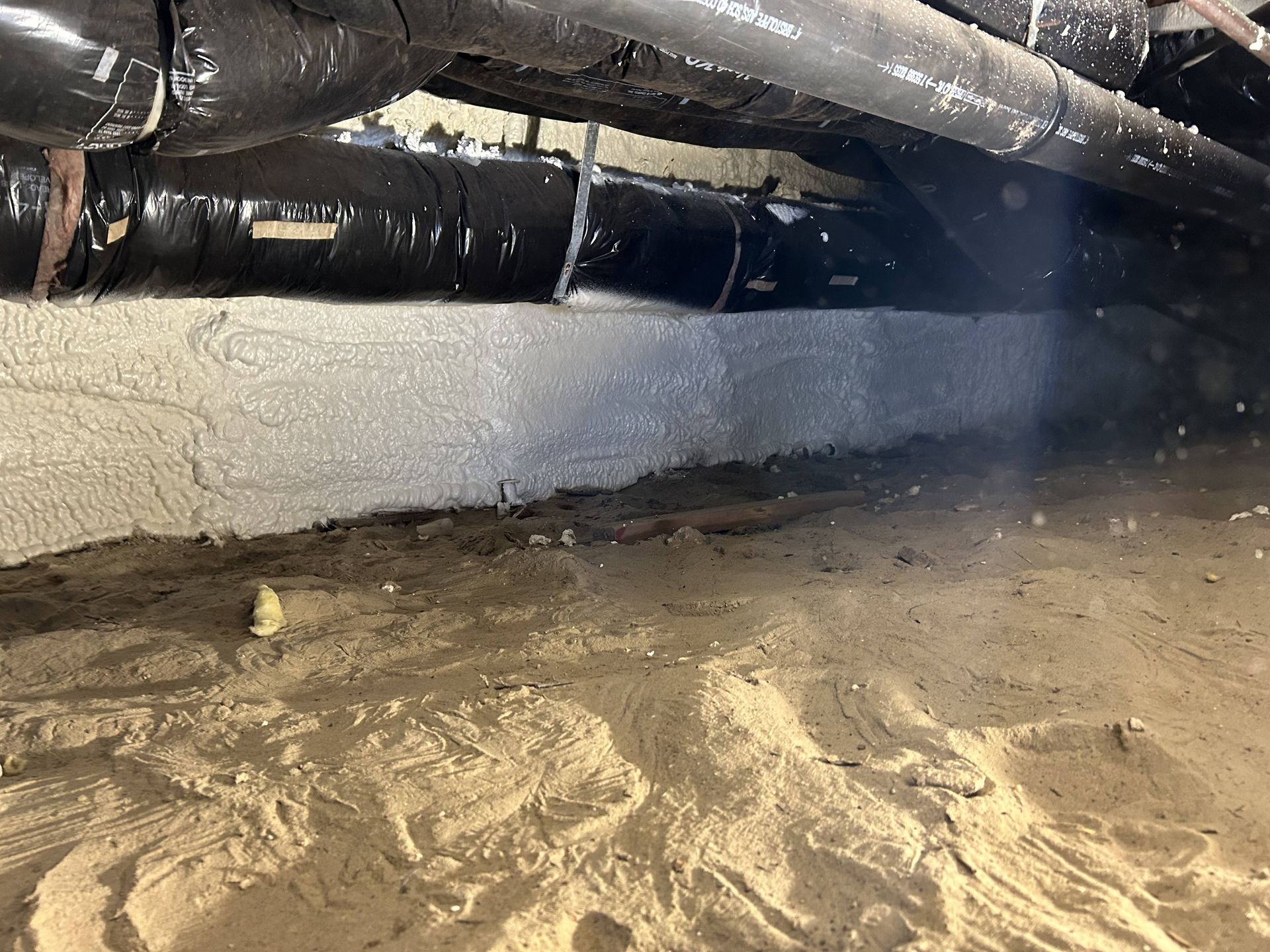 Basement crawlspace with insulated pipes overhead and a dirt floor, showing moisture or water damage.