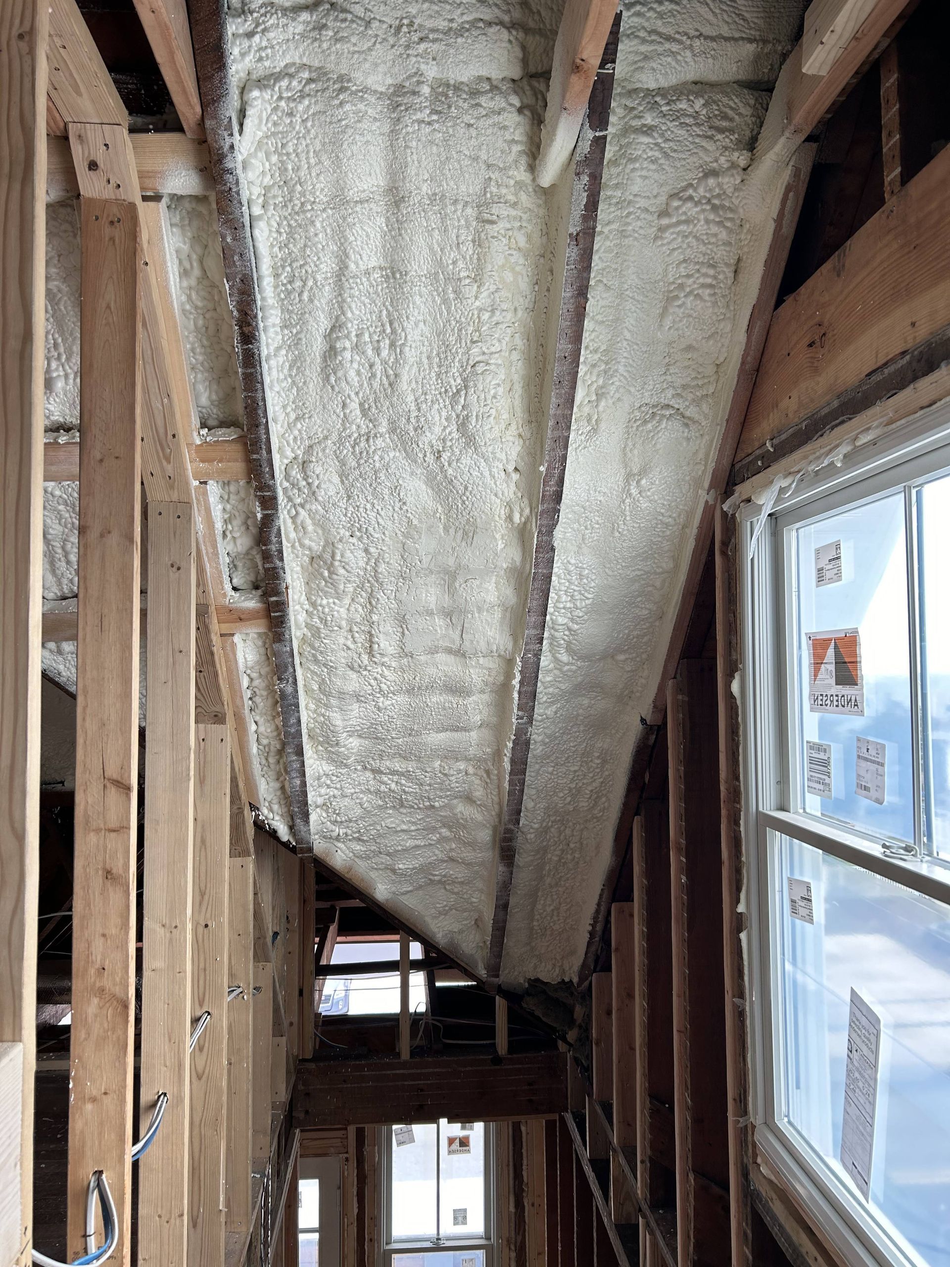 Foam-insulated ductwork running through an unfinished attic with exposed wooden framing and windows below