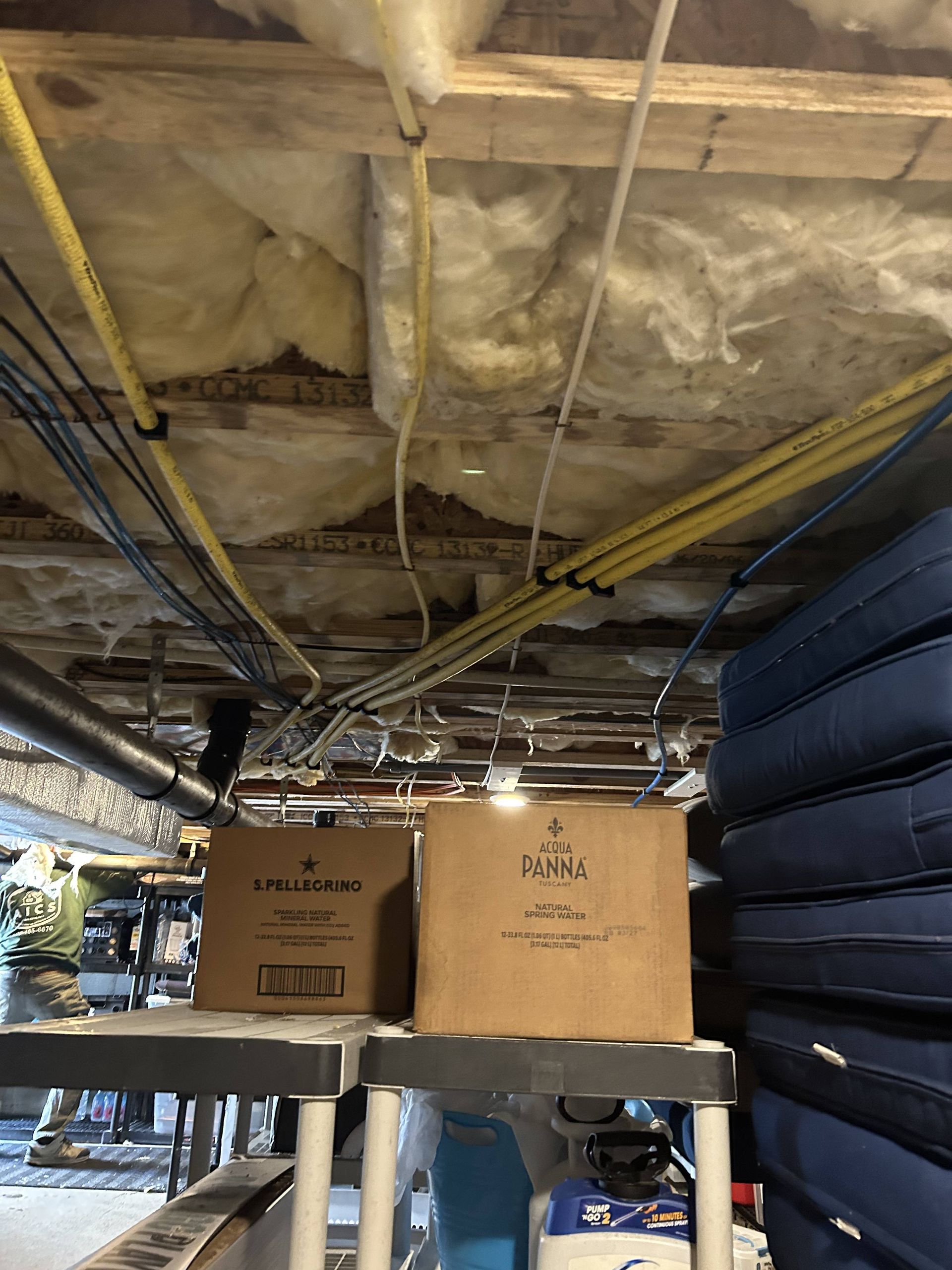 Storage area under a ceiling with exposed insulation, pipes, and cardboard boxes on a shelf.