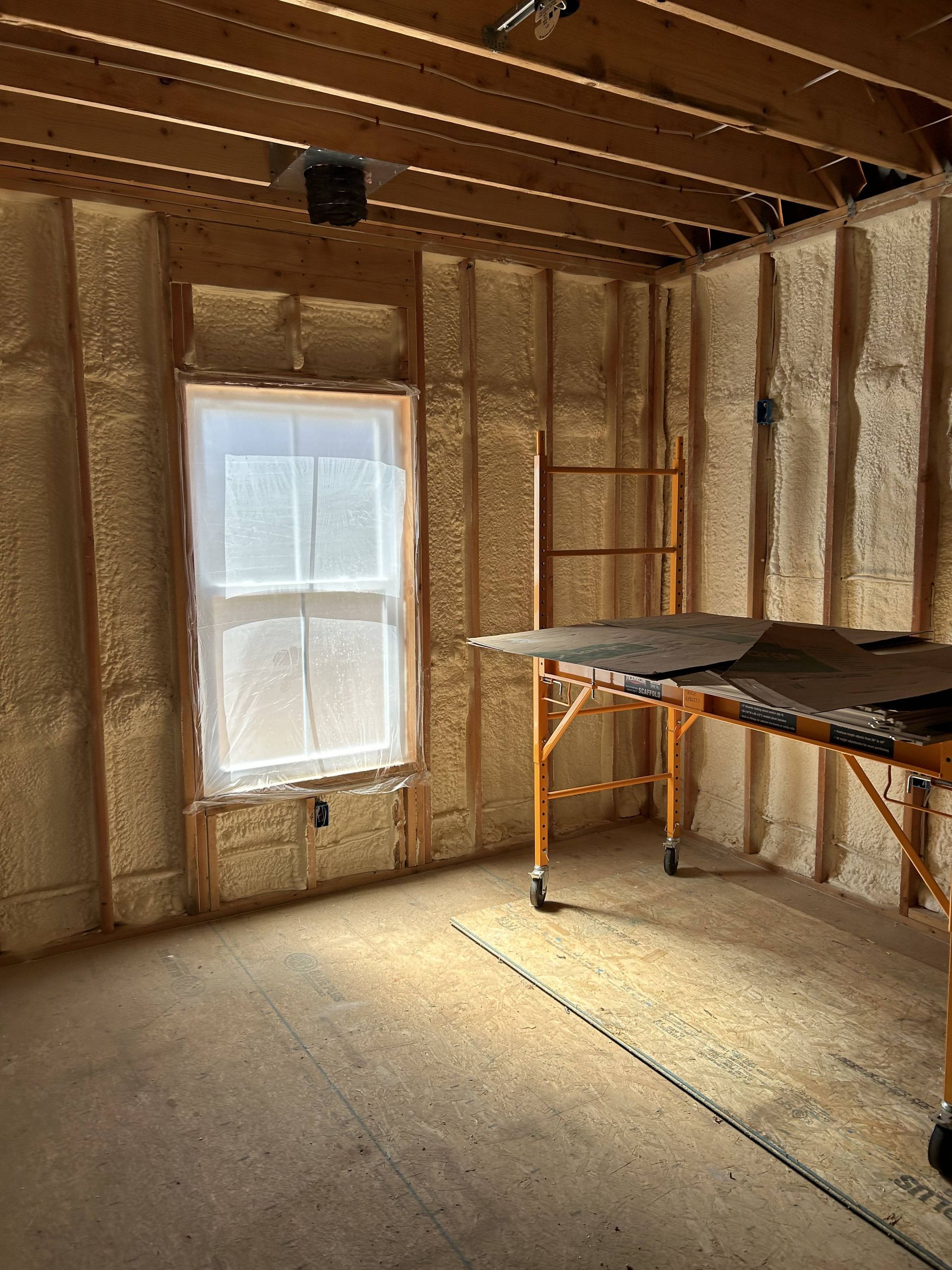 Unfinished room with insulated walls, exposed ceiling beams, a window, and a worktable under construction.